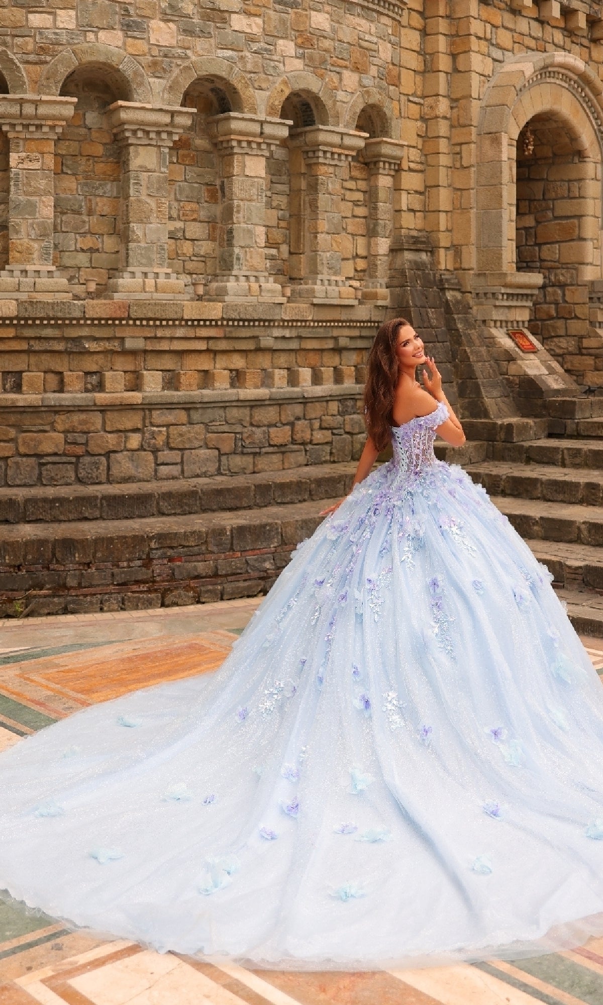 Wearing the Amarra 54231 3D Floral Quinceañera Dress, a woman stands outdoors before a stone building, glancing over her shoulder.